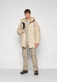 Puff jakke i lys beige, med hette, justerbare snorene og frontlommer. Bært med en grønn t-skjorte og beige cargo-bukser.