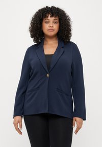 Blazer bleu marine en tissu doux, doté d'un bouton unique, de deux poches frontales et d'une coupe décontractée avec des manches longues.
