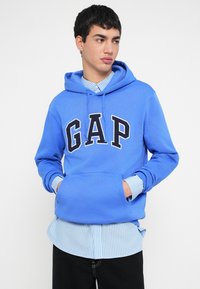 Modra jopa s kapuco z na tiskanim napisom "GAP" v mornarsko modri barvi, ki ima sprednji žep in vrvico za zategovanje kape, oblečena preko svetlo modre srajce z vzdolžnimi črtami. Črne hlače.