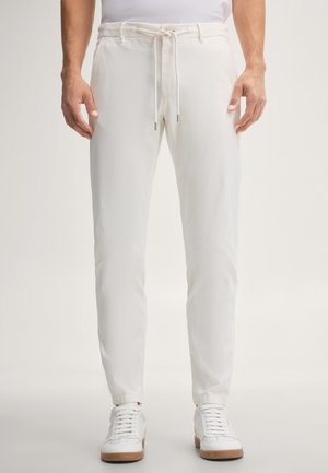 Man draagt slanke witte broek met aantrekkoord en witte sneakers, staand tegen een effen achtergrond.