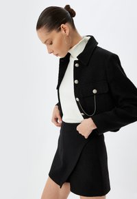 Veste noire texturée avec des boutons argentés et un accent en chaîne, portée sur un col roulé blanc à côtes et une jupe mini noire assortie.