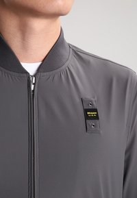 Gray bomber jacket with a smooth texture, ribbed collar, front zipper, and a branded black patch with yellow text on the left side.