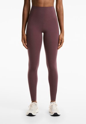 Persoon in hoog-geknipte mauve legging en witte sneakers, staande tegen een effen witte achtergrond.