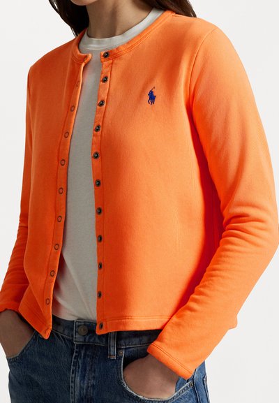 Orange ribbed cardigan with long sleeves, round neckline, button front, and a blue embroidered logo. Worn over a gray shirt and denim jeans.