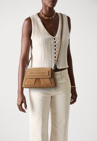 Borsa a tracolla in suede marrone con un design geometrico sulla patta e marchio impresso, abbinata a pantaloni a gamba larga di colore chiaro e un top in maglia.