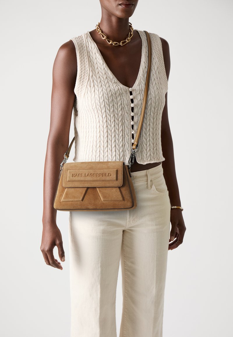 Borsa a tracolla in suede marrone con un design geometrico sulla patta e marchio impresso, abbinata a pantaloni a gamba larga di colore chiaro e un top in maglia.