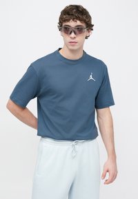 Σκούρο μπλε βαμβακερό T-shirt με στρογγυλή λαιμόκοψη και λευκό λογότυπο Jordan. Το μοντέλο φορά ανοιχτό μπλε φόρμα και αθλητικά γυαλιά ηλίου.