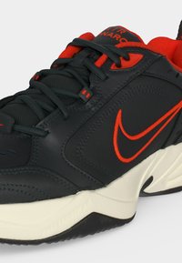 Baskets Nike Air Monarch noires et rouges avec lacets noirs, logo rouge et semelle blanche, présentées en gros plan en angle sur fond blanc.