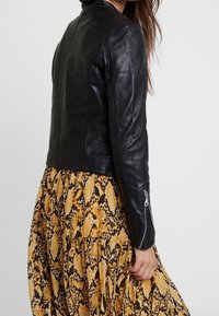 Giacca di pelle nera con un design aderente, aperture laterali con cerniera e una texture liscia, abbinata a un vestito con stampa di serpente marrone e gialla.