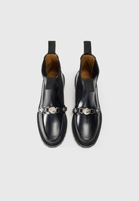 Bottines en cuir noir à enfiler avec des accents décoratifs argentés et des languettes de tirage sur fond blanc.