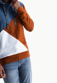 Sudadera con capucha en bloques de color, con secciones en azul, naranja y blanco, con cordones ajustables y una textura suave. Coordinada con vaqueros.