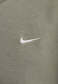 Tessuto a maglia verde con una texture sottile. Presenta un logo Nike ricamato bianco sul lato sinistro. Design semplice e lineare.