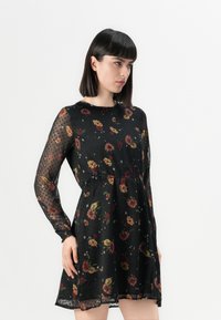 Robe florale noire avec une texture transparente à pois, des manches longues et une taille élastique. Motifs de fleurs multicolores avec de petits accents.