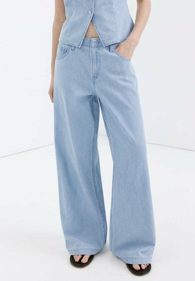 Personne portant un jean large bleu clair avec les mains dans les poches, associé à un top en denim sans manches assorti et des sandales noires.