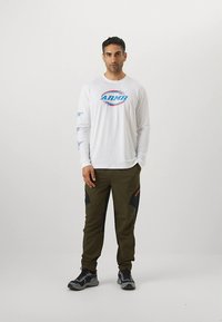 Biela košeľa s dlhým rukávom s modrým logom, skombinovaná so zelenými nohavicami olivovej farby s čiernymi panelmi a červenými detailmi; sivé topánky.