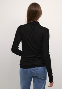 Top lungo nero a maniche lunghe con colletto alto, realizzato in tessuto leggero. La vista posteriore mostra una silhouette aderente e una leggera arricciatura all'orlo.