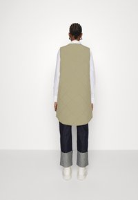 Lange, licht olijfgroene gewatteerde vest met een afgeronde zoom, gedragen over een wit shirt en gecombineerd met donkere jeans en witte sneakers.