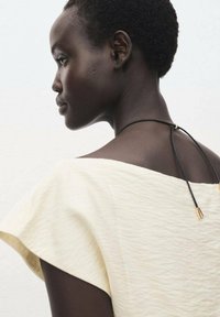 Haut texturé de couleur claire avec un large col, doté d'une fermeture à cordon réglable noir ornée d'un accent doré à l'arrière.