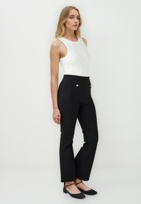 Zwarte high-waisted broeken met voorkruiszakken en gouden knopen, gecombineerd met een getailleerde witte tanktop. Model staat in een neutrale pose.