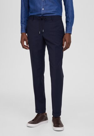 Pantalones azul marino con cintura ajustable y puños elásticos, combinados con zapatillas marrones con suelas blancas. Modelo lleva una camisa de mezclilla.