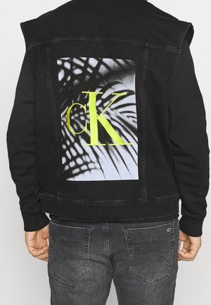 Osoba ubrana w czarną kurtkę dżinsową z dużym czarno-białym abstrakcyjnym wzorem liścia palmy oraz neonowożółtym logo "cK" na plecach.