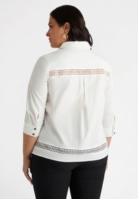 Blusa bianca con maniche a tre quarti, caratterizzata da un motivo in pizzo lungo la schiena e l'orlo. Tessuto morbido con dettagli in bottone sulle maniche.