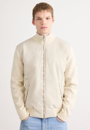 Ung man med kort hår, klädd i en beige zip-up jacka med ribbat krage och ärmar, blå jeans, stående mot en vit bakgrund.