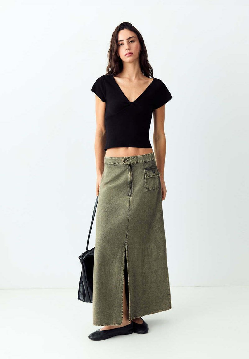 Jeune femme aux longs cheveux foncés portant un haut court noir à col en V, une longue jupe vert olive avec une fente à l'avant, des chaussures plates noires, et portant un sac noir.