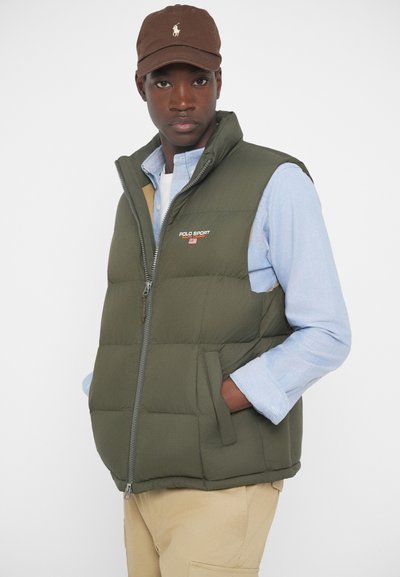 Zeleni pufasti jopič z zadrgo, stranskimi žepi in logotipom. Obut nad svetlo modro srajco, v paru z rjavim bejzbolskim klobukom.