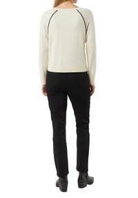 Frau steht mit dem Rücken zur Kamera, trägt ein cremefarbenes langärmeliges Oberteil mit schwarzen Nahtdetails, schwarze Jeans und schwarze Stiefeletten vor weißem Hintergrund.