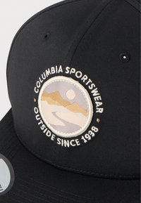 Μαύρο καπέλο με στρογγυλό αυτοκόλλητο που απεικονίζει κεντημένα βουνά και ήλιο. Ο σχεδιασμός περιλαμβάνει το κείμενο «Columbia Sportswear» και «Outside Since 1938».