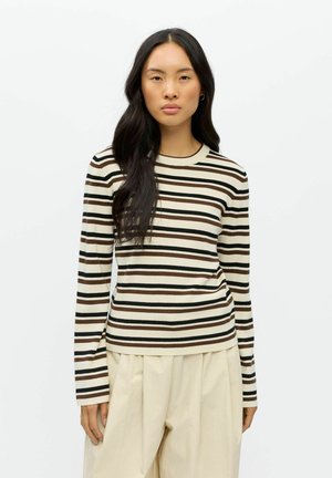 Femme aux longs cheveux foncés portant un haut à manches longues rayé crème, marron et noir, et un pantalon plissé beige clair, debout devant un fond blanc.