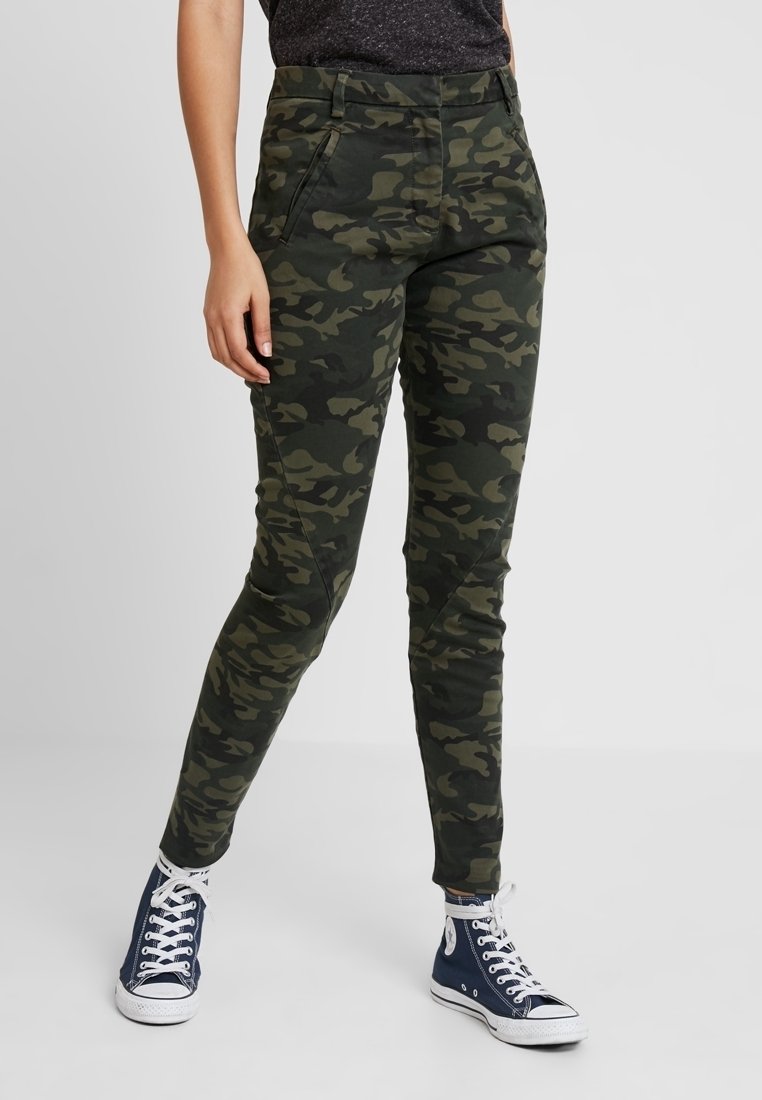 Maskáčové kalhoty v tmavě zelené a černé barvě, tapered střih s bočními kapsami. Spárovány s námořnickými teniskami s bílými detaily a tkaničkami.