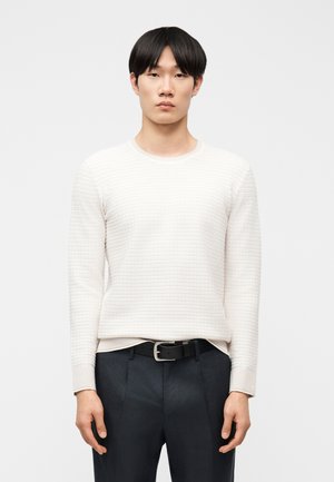 Mladý muž s rovnými čiernymi vlasmi, oblečený v textúrovanom bielom dlhorukáve svetri, ktorý má zastrčený do čiernych nohavíc s čiernym opaskom, stojaci pred bielym pozadím.