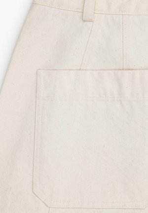 Zbliżenie na kremową kieszeń z tkaniny z widocznymi szwami i szlufką na pasek powyżej na teksturowanym materiale.