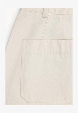 Zbliżenie na kremową kieszeń z tkaniny z widocznymi szwami i szlufką na pasek powyżej na teksturowanym materiale.