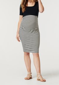 Kobieta w ciąży w czarnej bluzce i spódnicy ciążowej w czarno-białe paski sięgającej do kolan, w beżowych sandałach, stojąca na białym tle.