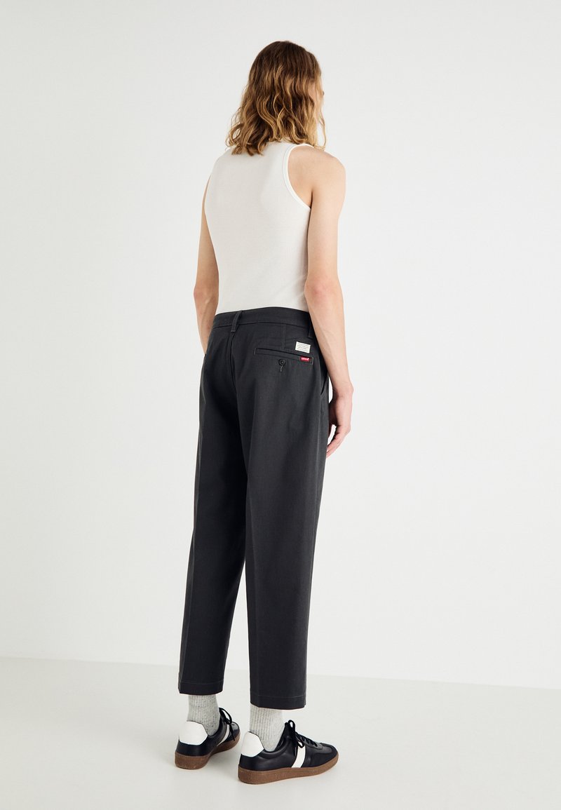 Pantalones grises de talle alto y corte recto. Hechos de una tela texturizada, con bolsillos traseros y una etiqueta de marca. Combinados con zapatos negros.