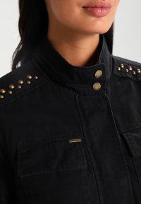 Černá bunda se strukturovaným límcem, zlatými hroty na ramenech a dvěma předními kapsami. Bavlněná tkanina s hladkou texturou.