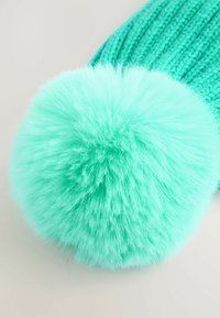 Bonnet tricoté turquoise vif avec un large pompon duveteux sur le dessus, présenté en gros plan sur un fond clair.