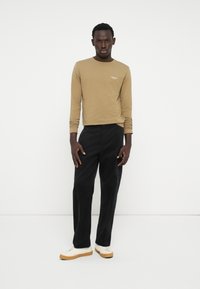 Camicetta in khaki a maniche lunghe, pantaloni in corduroy neri e sneakers bianche. La camicia presenta un piccolo logo sul petto. Design semplice e moderno.