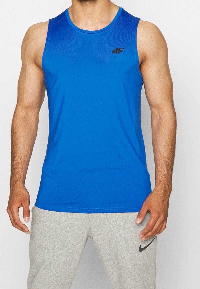 Hombre musculoso vestido con una camiseta deportiva sin mangas azul con logo y pantalones de chándal gris claro de Nike, de pie frente a un fondo sencillo.