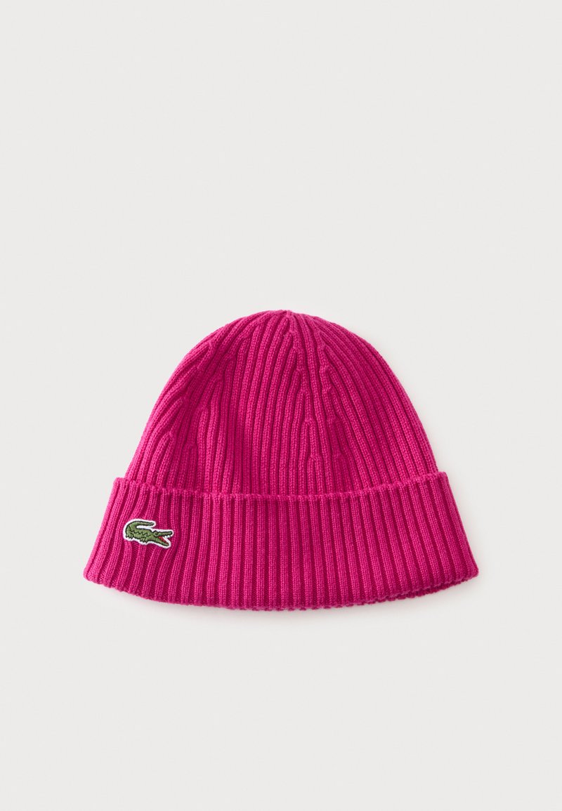 Bonnet en tricot côtelé rose vif avec revers, avec un petit logo crocodile vert sur le côté, sur un fond clair uni.