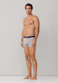 Mężczyzna bez koszulki, stojący boso, ubrany w szare bokserki z granatową gumą w pasie z napisem "Hackett", na jednolitym jasnoszarym tle.