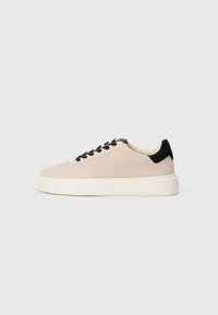 Beige semsket sneakers med sorte detaljer, rund tå, sorte snørebånd, og en kontrasterende hvit gummisåle. Minimalistisk design, glatt tekstur.