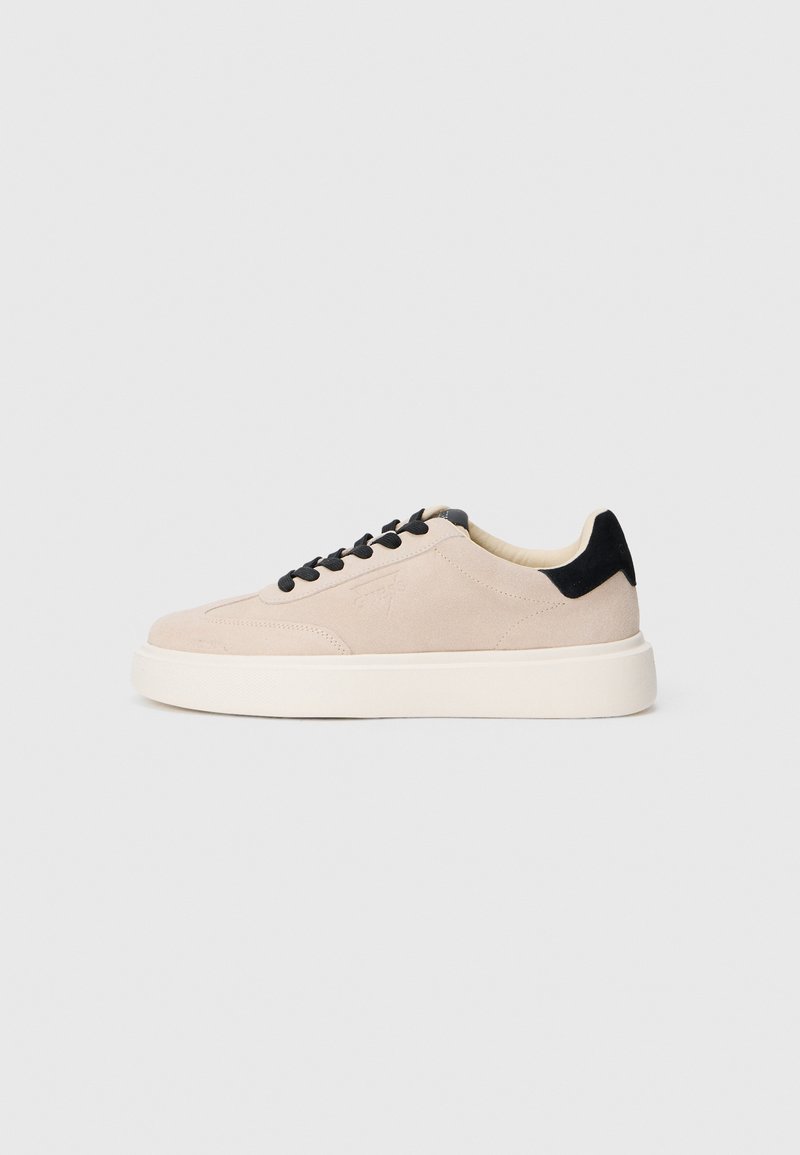 Beige semsket sneakers med sorte detaljer, rund tå, sorte snørebånd, og en kontrasterende hvit gummisåle. Minimalistisk design, glatt tekstur.