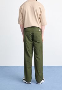Beige kortærmet skjorte, olivenfarvede bukser med en afslappet pasform, synlige baglommer og et logomærke på den højre baglomme. Sorte og hvide sneakers.
