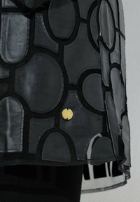 Schwarzes Lederkleidungsstück mit einem geometrischen Muster aus Formen, an dem ein goldener Anhänger befestigt ist. Das Design verfügt über eine strukturierte Oberfläche.