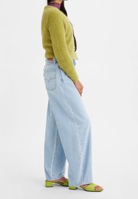 Zelený huňatý kardigan, světle modré volné džíny a zelené sandály na podpatku. Významné textury zahrnují jemnou tkaninu a denim. Jednoduchý design.