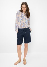 Blusa leggera a maniche lunghe con motivo floreale, posizionata su uno sfondo neutro; abbinata a pantaloni corti blu navy e sandali color sabbia per un look informale.